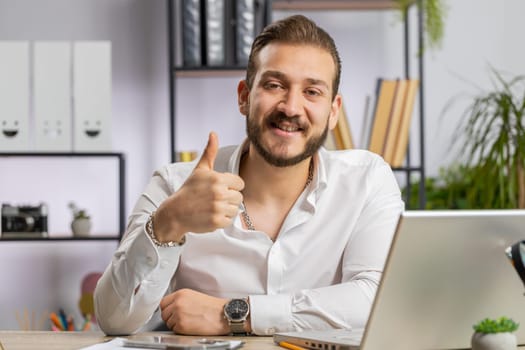 Like. Happy middle eastern businessman guy working on laptop looking approvingly at camera showing thumbs up, like sign positive something good, positive feedback at office workplace. Freelancer man
