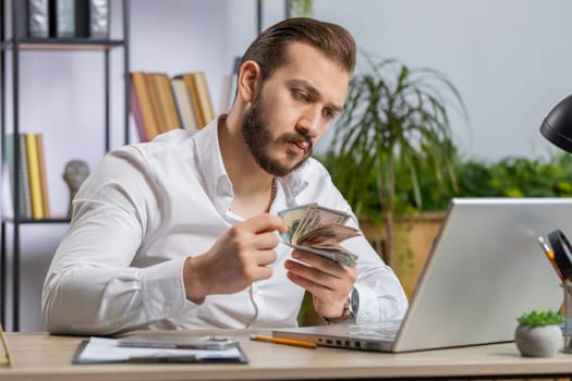 Rich middle eastern businessman working on laptop pc counting money cash, calculate earnings income profit at home office workplace. Professional freelancer man. Business people. Employment occupation