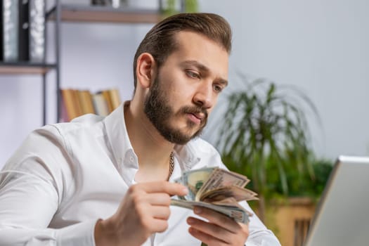 Rich middle eastern businessman working on laptop pc counting money cash, calculate earnings income profit at home office workplace. Professional freelancer man. Business people. Employment occupation