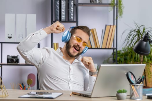 Happy relaxed overjoyed middle eastern businessman working on laptop computer at office wearing headphones listening favorite energetic disco music and dancing. Freelancer man relaxing, taking a break