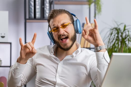 Happy relaxed overjoyed middle eastern businessman working on laptop computer at office wearing headphones listening favorite energetic disco music and dancing. Freelancer man relaxing, taking a break
