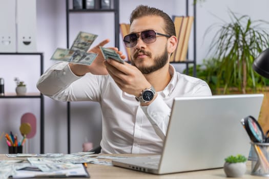 Cheerful rich young lebanese businessman working on laptop, wasting throwing money to camera more tips earnings big profit success win lottery shopping. Happy freelancer arab man at home office desk
