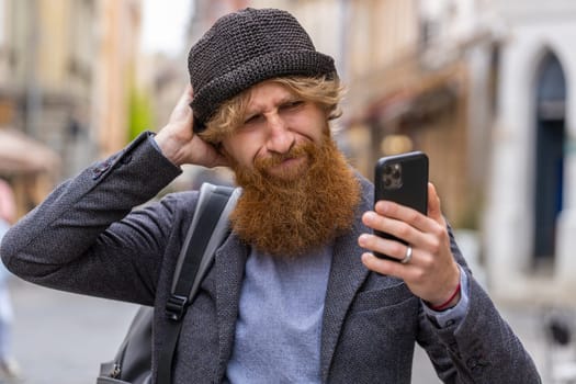 Surprised bearded man use smartphone typing browsing, loses becoming surprised sudden lottery results, bad news, fortune loss, fail. Redhead guy walking in urban city street outdoors. Town lifestyles