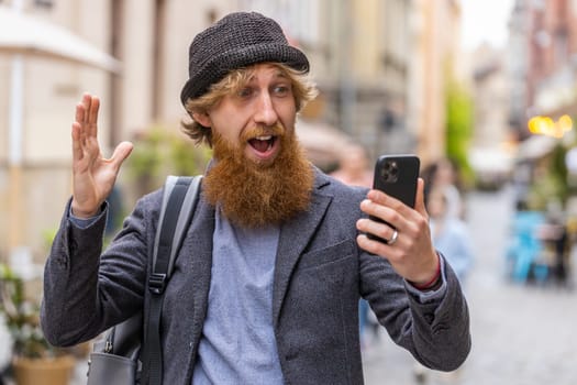 Oh my God Wow. Excited happy joyful winner young adult man use smartphone typing browsing celebrating win good message news. Redhead bearded guy with mobile phone walking in urban city street outdoors