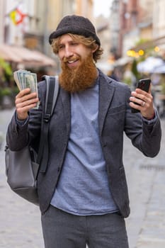 Happy young bearded man counting money dollar cash, use smartphone calculator app, satisfied of income for planned vacation gifts. Redhead guy walking in urban city street. Outdoors. Vertical shot