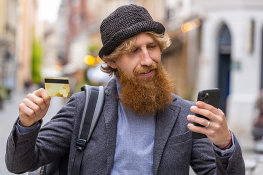 Young bearded man using credit bank card smartphone while transferring money, purchases online shopping, order food delivery, booking hotel room. Redhead guy walking in urban city street outdoors