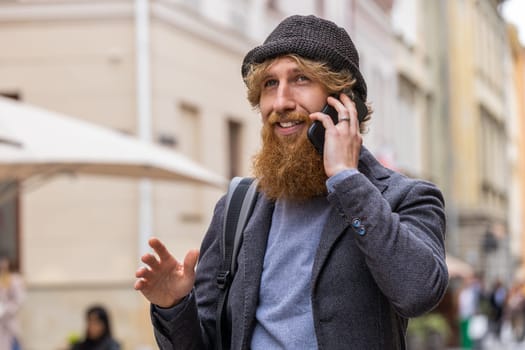 Happy bearded man having remote conversation communicate speaking by smartphone. Handsome young adult guy talking on phone unexpected good news gossip walking in city street. Town lifestyles outdoors