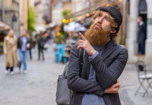 Thoughtful pensive puzzled man with pensive expression, pondering a solution, doubting question, thinking inspiration dreaming outdoors. Redhead guy in city street pointing empty place. Copy-space