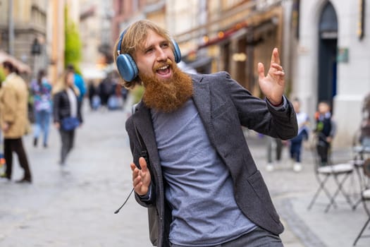 Happy overjoyed young adult man in wireless headphones listening favorite energetic music, celebrating win. Redhead bearded guy tourist traveler walking in urban city street. Town lifestyles outdoors