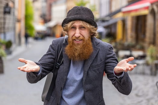 What. Why. Surprised frustrated young bearded man raising hands asking reason of failure, demonstrating disbelief irritation by troubles outdoors. Confused redhead guy walking in urban city street
