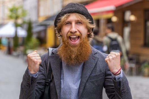Portrait of young man shouting, celebrating success, winning, goal achievement, good news, lottery jackpot luck, victory outdoors. Redhead bearded guy walking in urban city street. Town lifestyles
