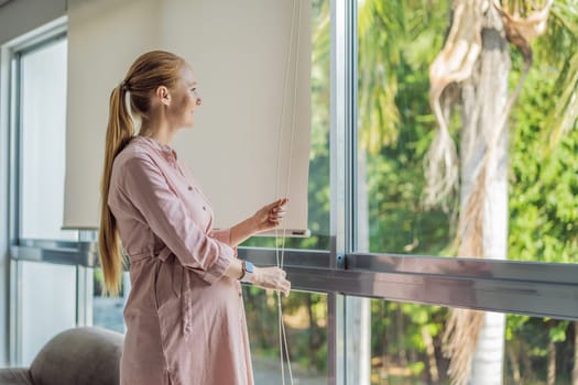 Portrait of young pregnant woman standing near window holding hands on her belly at modern home with copy space. Pregnancy, motherhood, expectation and tenderness concept.