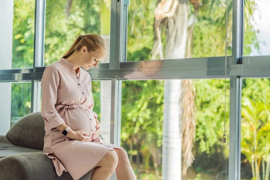 Portrait of young pregnant woman sitting near window holding hands on her belly at modern home with copy space. Pregnancy, motherhood, expectation and tenderness concept.