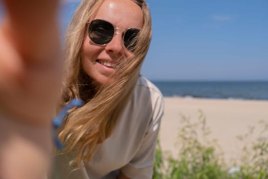 Young woman in sunglasses with shell bracelets looking into camera. Influencer Using mobile phone for video call selfie waving on the beach seacoast. Holiday vacation advertisement concept. Social media