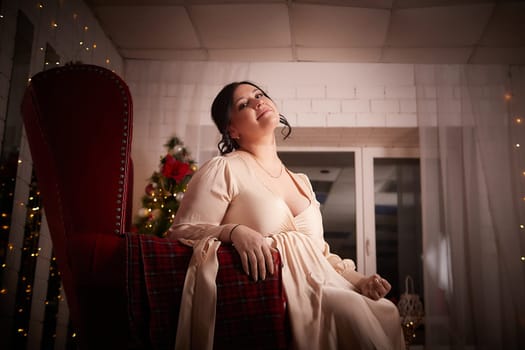 Owerweight elegant Woman at Christmas room. Fat plumb pretty girl in a beautiful dress for a holiday. Buxom female model posing alone in New year sudio