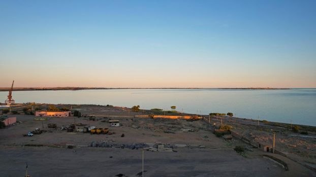 Abandoned territory on the beach of Lake Balkhash. The gradient of the sky at sunset. There is a crane and old fences, buildings. People are walking on the beach. Reeds grow in the water. Kazakhstan