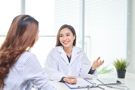 Female doctor talks to female patient in hospital office while writing on the patients health record on the table. Healthcare and medical service. 