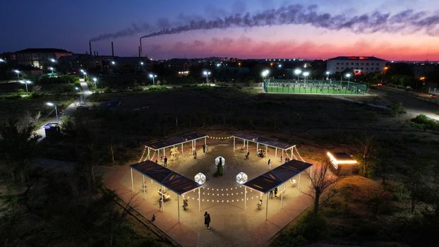 People are relaxing in the evening park. Lanterns and garlands are lit. Children ride on a swing. Beautiful lighting of the recreation area. There are sports grounds in the distance. Green trees.