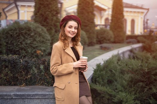 Beautiful smiling Parisian woman holding a paper cup of fresh coffee standing outdoors beautiful garden posing for female fashion. Portrait of stylish young woman wearing autumn coat and red beret.