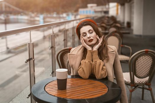 Parisian woman with coffee mug on city street in evening. Charming woman enjoying her cup of coffee sitting in a spring outdoor cafe. French woman in red beret with background of urban city.