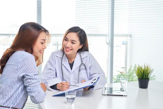 Female doctor talks to female patient in hospital office while writing on the patients health record on the table. Healthcare and medical service. 