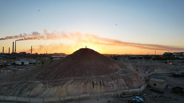 Two silhouettes of guys on the mountain. Behind them, the factory smokes at the sunset of the orange sun. An epic moment. Abandoned garages. The threat to the environment. Balkhash, Kazakhstan