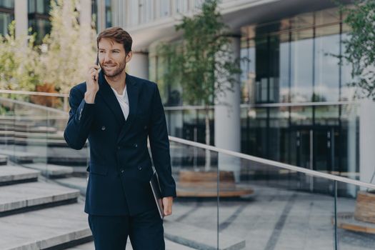 Fashionable businessman uses modern technologies has telephone conversation holds electronic gadget poses outdoor near office center. Successful economist makes phone call focused into distance