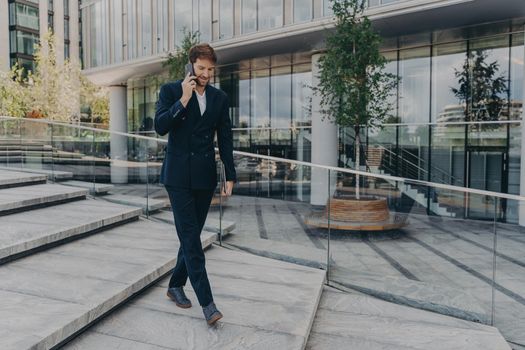 Happy caucasian businessman talking on smartphone while walking along office center with laptop computer in hand. Young successful entrepreneur having friendly conversation outdoors in urban cityscape