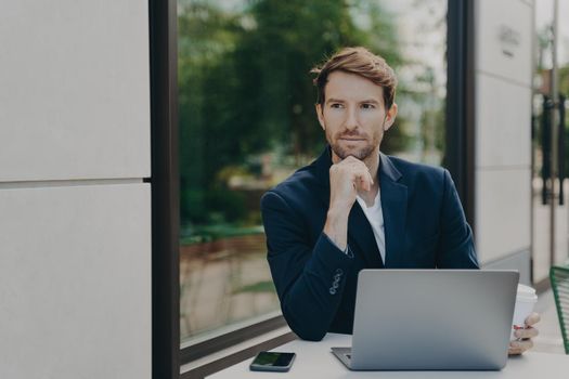 Male ceo with thoughtful expression browses fast wireless mobile internet works on laptop computer in street cafe drinks takeaway coffee surrounded by modern gadgets prepares business project