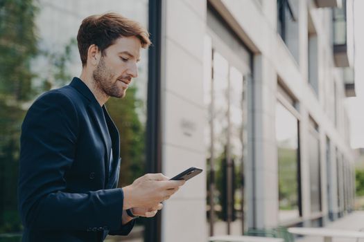 Focused handsome man with beard wearing dark suit chatting on smartphone, browsing web on phone while standing in front of building with lots of windows, selective focus on businessman typing message