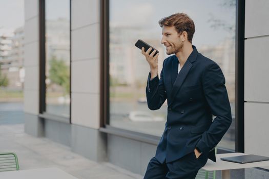 Confident businessman with smartphone using voice assistant app to organize his day or recording message, speaking on speakerphone, standing outside with hand in pocket while leaning on cafe table
