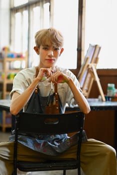 Young hipster queer artist holding palette and brush sitting in front of easel in bright studio. Art, education, creativity concept.