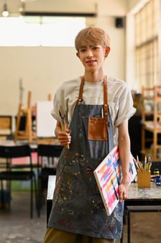 Young creative queer artist holding palette and brush standing in bright studio. Art, education, creativity and people concept.