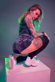 Beautiful young blonde tattoed girl in blue velour booty shorts and t-shirt, making squats on top of big cube, showing off her amazing booty