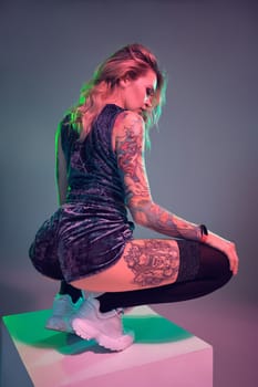 Beautiful young blonde tattoed girl in blue velour booty shorts and t-shirt, making squats on top of big cube, showing off her amazing booty