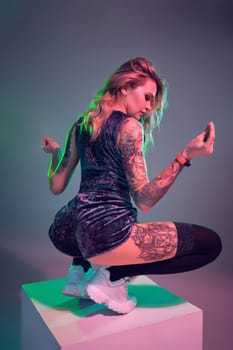 Beautiful young blonde tattoed girl in blue velour booty shorts and t-shirt, making squats on top of big cube, showing off her amazing booty