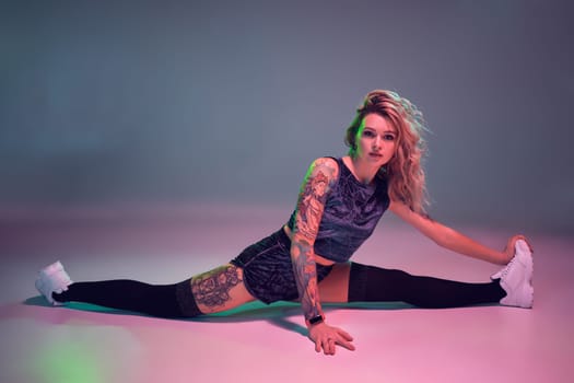 Young beautiful caucasian blonde girl with tattoo on her body, wearing velour blue booty shorts and black stockings making stretching before dancing twerk, she sits on the splits, pink background
