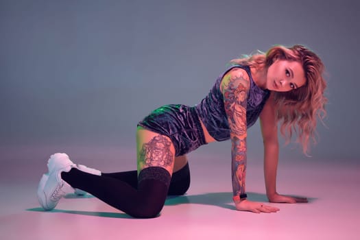 Young beautiful caucasian blonde girl with tattoo on her body, wearing velour blue booty shorts and black stockings dancing twerk on her knees, pink background
