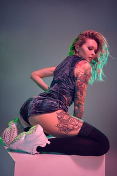 Beautiful young blonde tattoed girl in blue velour booty shorts and t-shirt, she sits on top of big cube, showing off her amazing booty from behind