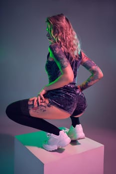 Beautiful young blonde tattoed girl in blue velour booty shorts and t-shirt, making squats on top of big cube, showing off her amazing booty