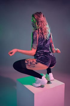 Beautiful young blonde tattoed girl in blue velour booty shorts and t-shirt, making squats on top of big cube, showing off her amazing booty