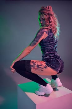 Beautiful young blonde tattoed girl in blue velour booty shorts and t-shirt, making squats on top of big cube, showing off her amazing booty