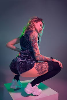 Beautiful young blonde tattoed girl in blue velour booty shorts and t-shirt, making squats on top of big cube, showing off her amazing booty