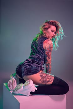Beautiful young blonde tattoed girl in blue velour booty shorts and t-shirt, she sits on top of big cube, showing off her amazing booty from behind