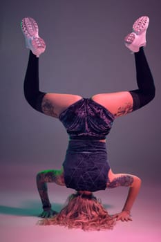 Young beautiful caucasian blonde gymnast woman with tattoo on her body, wearing velour blue booty shorts and black stockings standing upside down, pink background