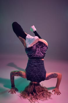Young beautiful caucasian blonde gymnast woman with tattoo on her body, wearing velour blue booty shorts and black stockings standing upside down, pink background