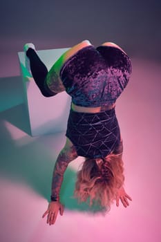 Young beautiful caucasian blonde gymnast woman with tattoo on her body, wearing velour blue booty shorts and black stockings standing upside down using big cube, pink background