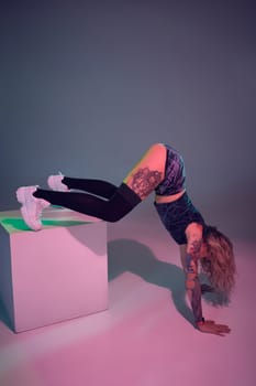 Young beautiful caucasian blonde gymnast woman with tattoo on her body, wearing velour blue booty shorts and black stockings standing upside down using big cube, pink background