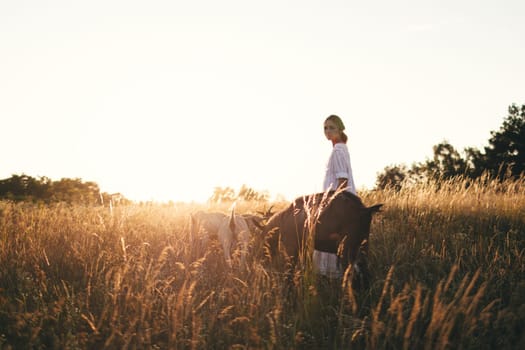 Young woman in white dress is walking with goats on the meadow at sunset. Attractive female farmer feeding her goats on her small business organic farm.