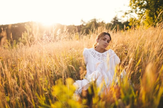 Beautiful girl in white dress sitting in wheat field at sunset